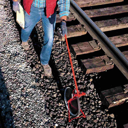 Person using a Rolatape measuring wheel along a railroad track, dressed in workwear and holding a clipboard.