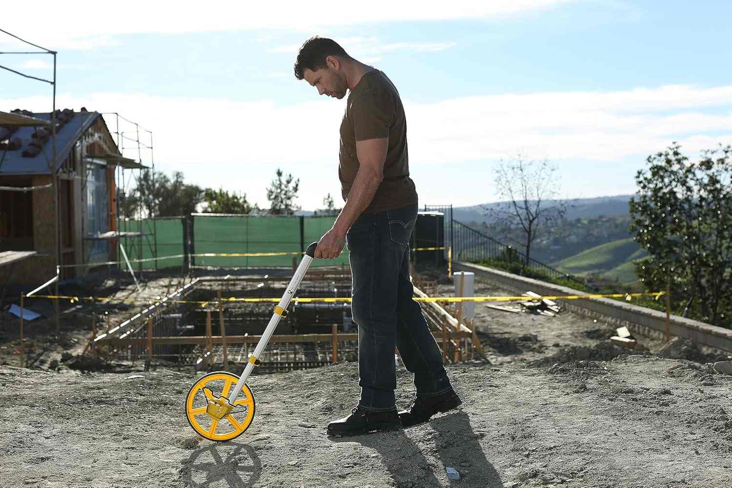 A landscaping professional walking with the Rolatape RT300 measuring wheel on uneven dirt terrain at a residential jobsite, highlighting the wheel's durability and the comfort of the ergonomic pistol grip.