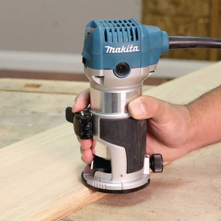 Close-up action shot of a woodworker using the Makita RT0701C corded compact woodworking router to smoothly shape the edge of a light wood board, highlighting the tool's ergonomic grip and fixed base.