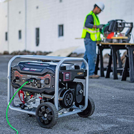 Lifestyle image showing the generator operating on a job site with a green power cord plugged in, while a worker operates a saw in the background.
