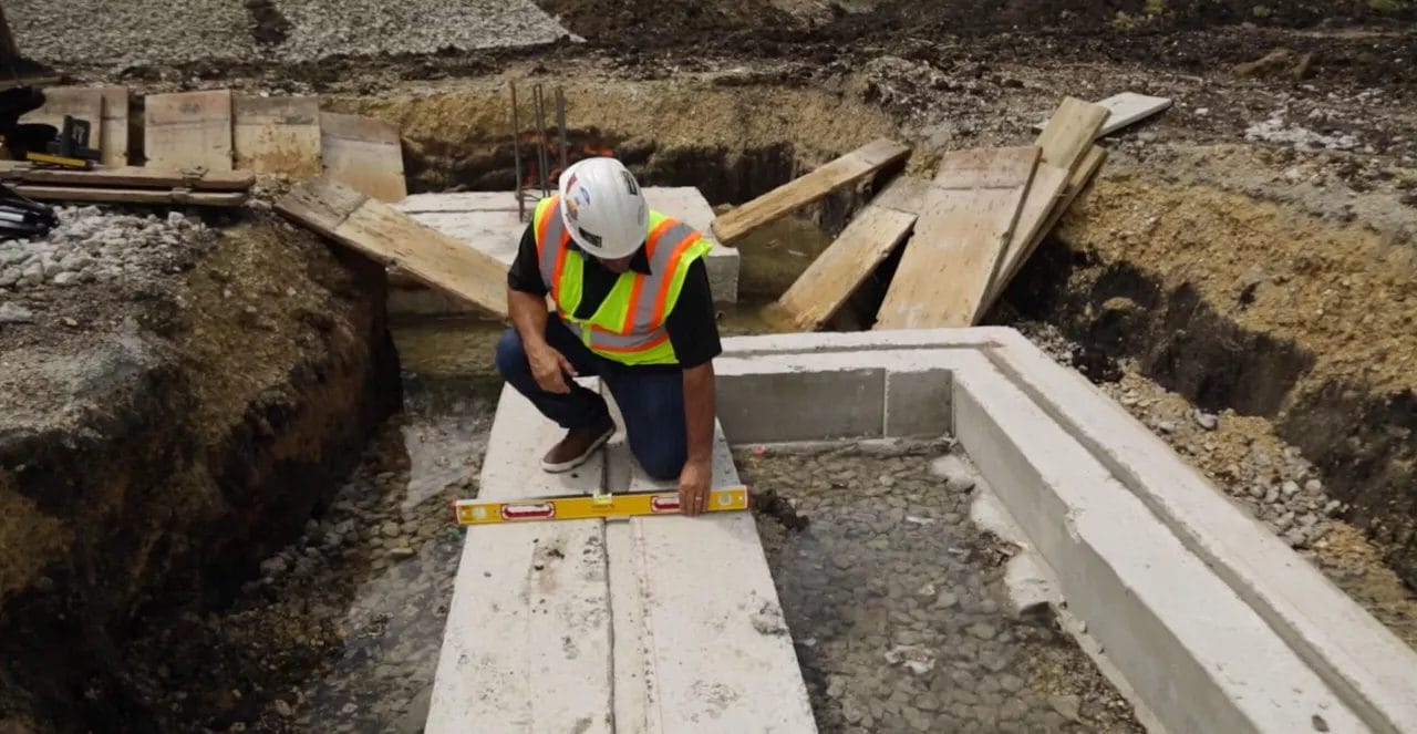 A construction worker kneeling on a concrete footing, using the yellow STABILA level horizontally to check the levelness of the surface during construction in a dug-out trench.