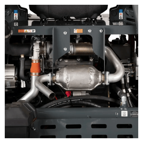 The image provides a close-up view of the engine compartment of a piece of heavy machinery, likely a concrete power trowel based on our recent conversation. You can see various mechanical components, including metal pipes and hoses, a corrugated exhaust component wrapped in insulation, and a larger, textured silver muffler or catalytic converter. There are also electrical wires and warning labels visible within the machinery's frame. The overall impression is of a complex and robust engine system.