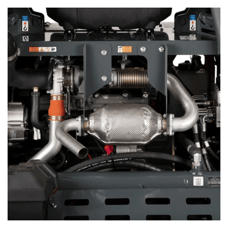 The image provides a close-up view of the engine compartment of a piece of heavy machinery, likely a concrete power trowel based on our recent conversation. You can see various mechanical components, including metal pipes and hoses, a corrugated exhaust component wrapped in insulation, and a larger, textured silver muffler or catalytic converter. There are also electrical wires and warning labels visible within the machinery's frame. The overall impression is of a complex and robust engine system.