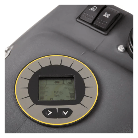 
The image shows a close-up of a digital display, likely from a piece of machinery. The display is round with a gray background and a yellow outline. It's showing a number "995 RPM" in the center, which likely indicates the revolutions per minute of some part of the machine. There are also other symbols and numbers around the edge of the display, including a symbol of a seat, possibly indicating the machine has a seat, and a headlight symbol. Below the display are two arrow buttons, one pointing left and on