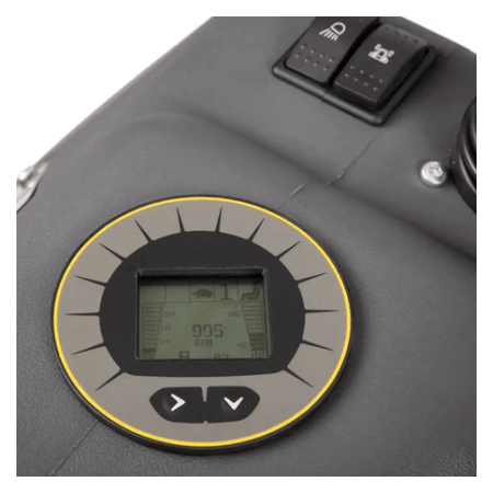 
The image shows a close-up of a digital display, likely from a piece of machinery. The display is round with a gray background and a yellow outline. It's showing a number "995 RPM" in the center, which likely indicates the revolutions per minute of some part of the machine. There are also other symbols and numbers around the edge of the display, including a symbol of a seat, possibly indicating the machine has a seat, and a headlight symbol. Below the display are two arrow buttons, one pointing left and on