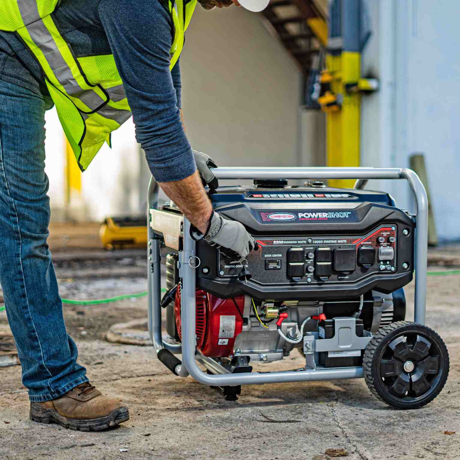 Lifestyle image of a construction worker wearing a safety vest and hard hat starting the generator on a job site with a green extension cord plugged in.