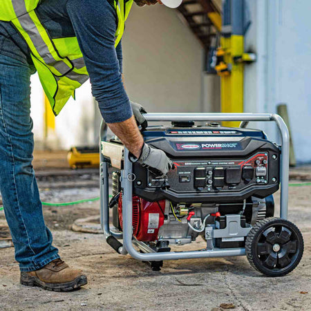 Lifestyle image of a construction worker wearing a safety vest and hard hat starting the generator on a job site with a green extension cord plugged in.