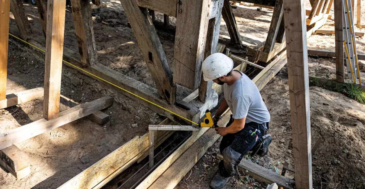 Worker using a yellow tape measure and square to align wooden beams at a construction site, demonstrating precise angle measurement and structural layout accuracy essential for framing and foundational carpentry work.