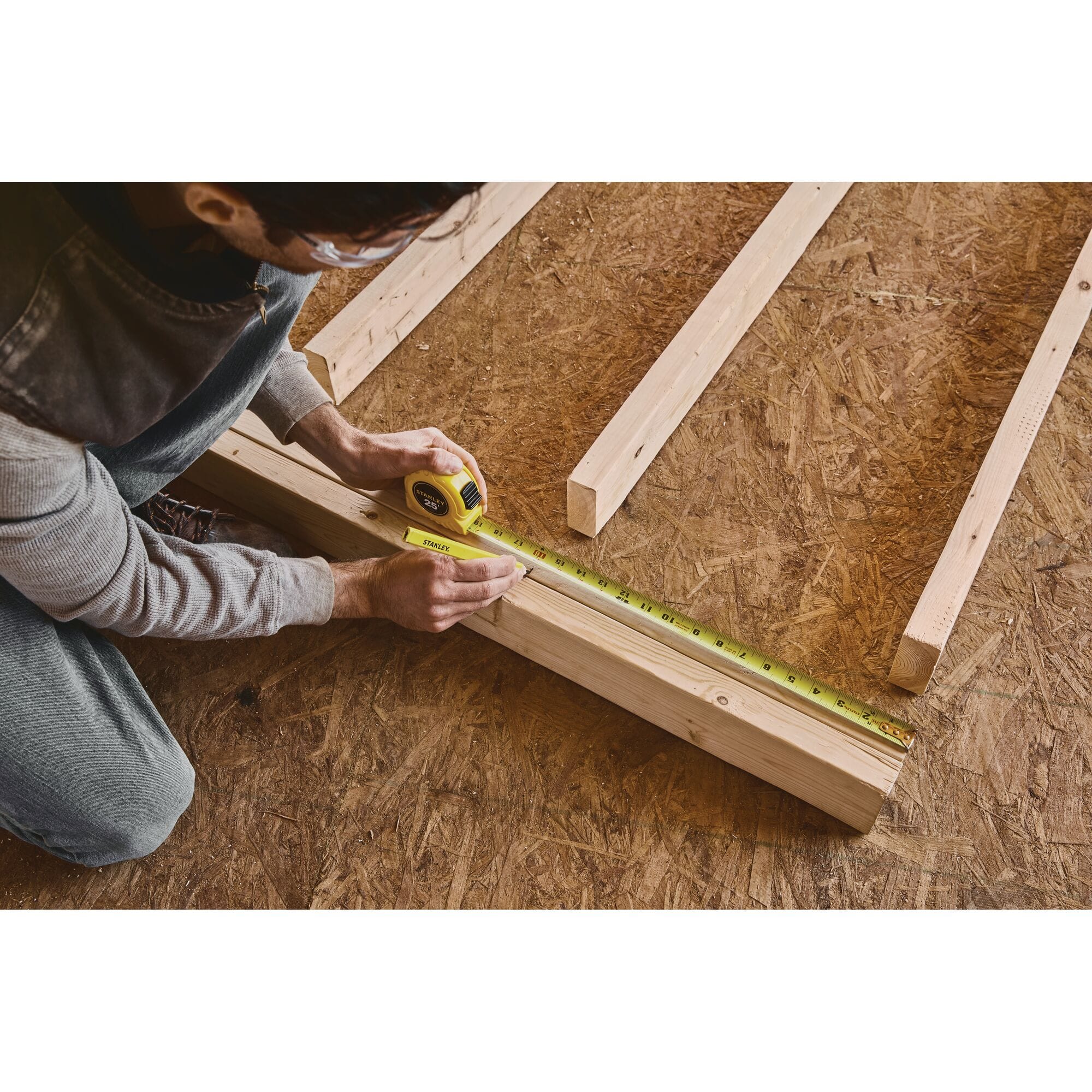A real-world residential construction application photograph of an overhead shot showing a worker kneeling on OSB subflooring, using the yellow Stanley 25-foot tape measure and a carpenter's pencil to mark layout lines on a wood framing board.