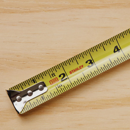 A top-down detailed graphic look at the yellow steel measuring blade ribbon of the Stanley PowerLock rule, displaying clean black standard numbers, fractional lines, red 25ft text indicator box, and the stamped Stanley brand name on a wood background.