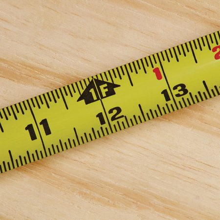 A macro close-up photograph tracking the yellow steel blade of the Stanley PowerLock 25-foot tape rule, focusing on the 12-inch milestone highlighted by a distinct black arrow graphic indicating one linear foot.