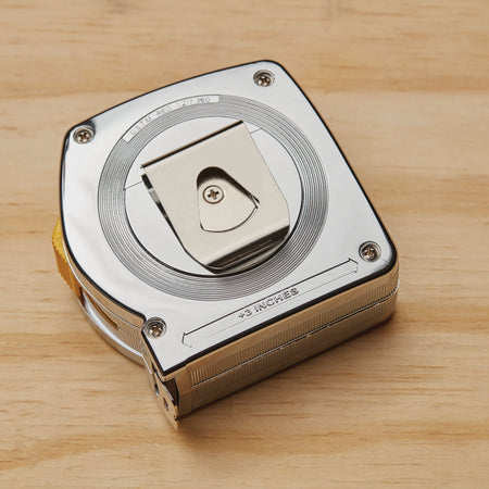 A flat-lay studio product shot displaying the reverse side of the polished chrome Stanley tape measure housing, showcasing the attached silver metal pocket clip, assembly screws, and the stamped technical casing measurement notation indicating plus three inches.