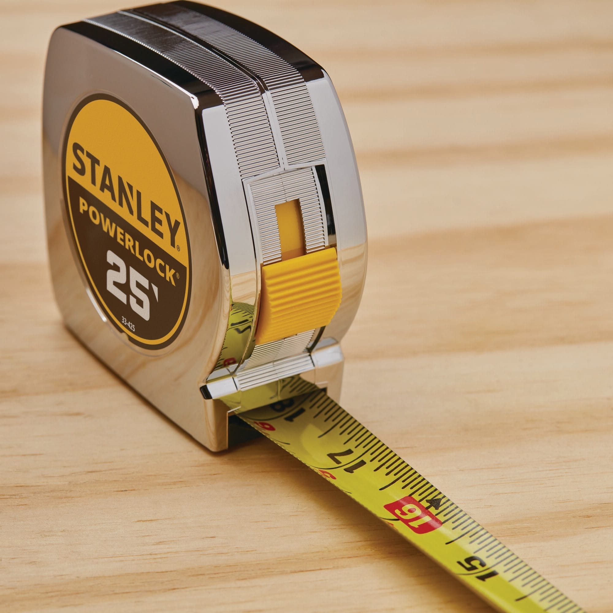 An angled foreground perspective view of the Stanley 33-425 tape rule resting on a light-colored wood surface, showcasing the yellow steel measuring ribbon extending past the 15-inch, 16-inch, and 17-inch fractional markings while displaying the bright yellow plastic slide lock button.