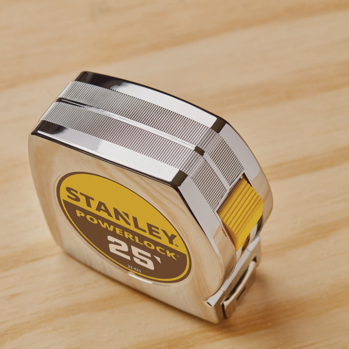 A high-angle studio shot looking down at the compact chrome case of the Stanley 25-foot PowerLock tool, highlighting the textured gripping lines etched into the metallic ABS shell and the recessed yellow thumb switch mechanism.