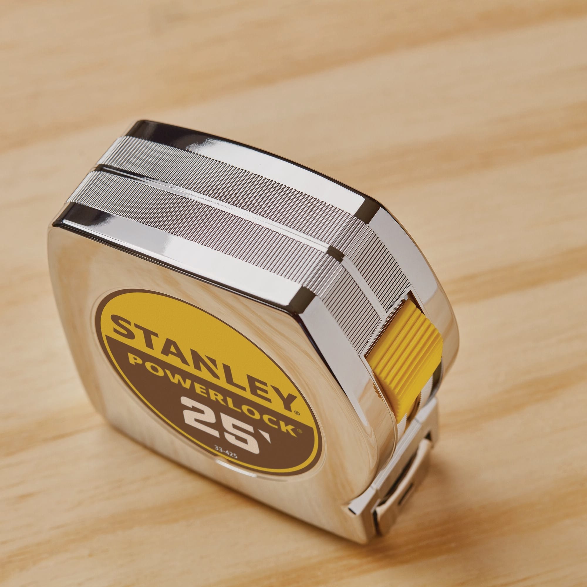 A high-angle studio shot looking down at the compact chrome case of the Stanley 25-foot PowerLock tool, highlighting the textured gripping lines etched into the metallic ABS shell and the recessed yellow thumb switch mechanism.