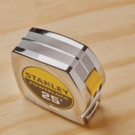A high-angle studio shot looking down at the compact chrome case of the Stanley 25-foot PowerLock tool, highlighting the textured gripping lines etched into the metallic ABS shell and the recessed yellow thumb switch mechanism.
