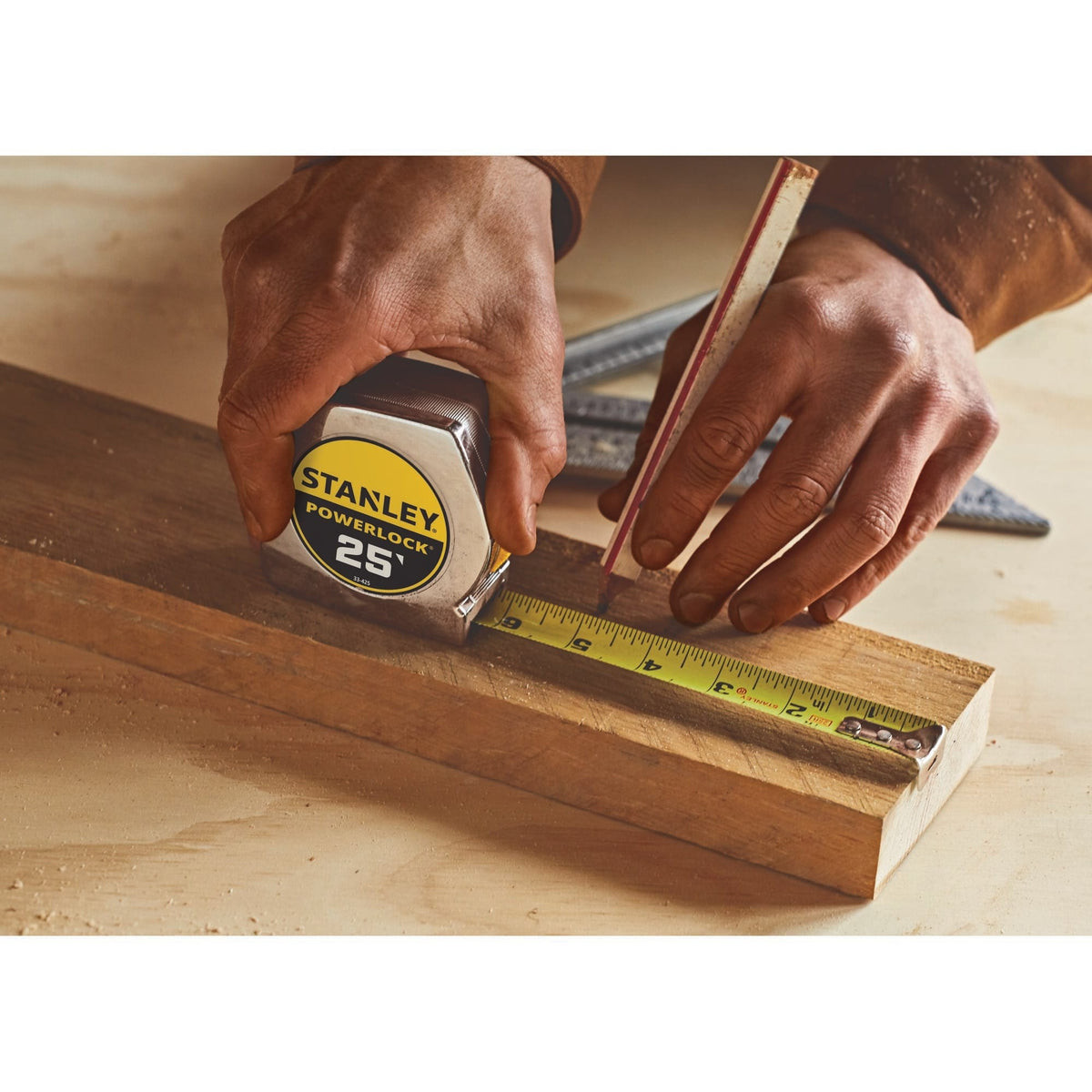 A jobsite lifestyle photograph focusing on a carpenter's hands using the chrome Stanley PowerLock 25-foot tape measure and a traditional timber pencil to accurately draw a dimension line onto a structural wood board.
