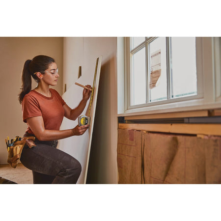 An indoor construction application shot showing a female tradesperson wearing protective safety glasses using the chrome Stanley 25' pocket rule to take a vertical alignment measurement on a drywall panel near a window frame.