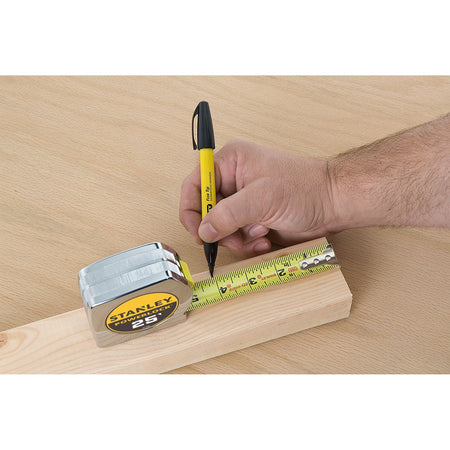 A close-up studio lifestyle photo of a technician using a yellow fine-tip permanent marker to trace a precise dimension tick mark along the extended high-contrast steel blade of the Stanley PowerLock tape rule over lumber.