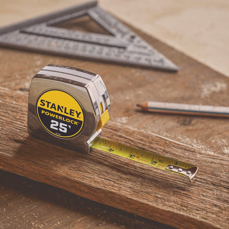 A close-up professional product shot of the Stanley PowerLock 25-foot tape measure resting face-forward on a rustic timber beam with its yellow blade slightly extended next to a carpenter's pencil and a metal speed square.