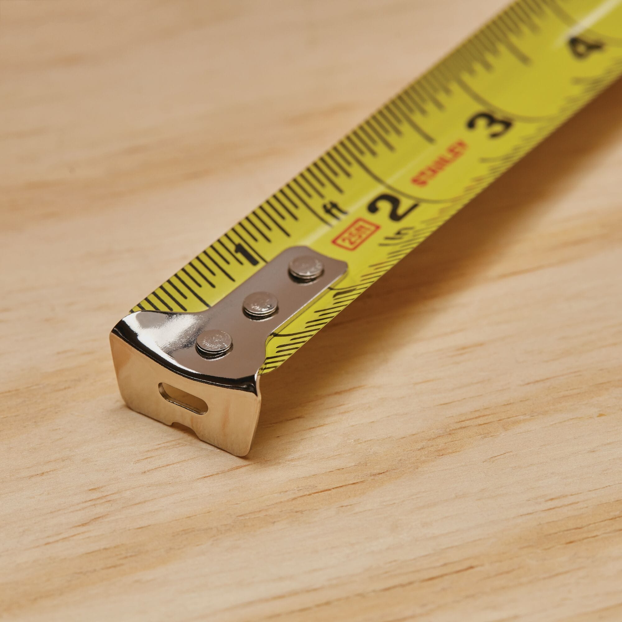 An extreme macro close-up photograph tracking the front tip of the extended yellow Stanley tape measure blade resting on a light plywood surface, highlighting the three-rivet configuration of its shiny chrome metal end hook.