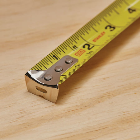 An extreme macro close-up photograph tracking the front tip of the extended yellow Stanley tape measure blade resting on a light plywood surface, highlighting the three-rivet configuration of its shiny chrome metal end hook.