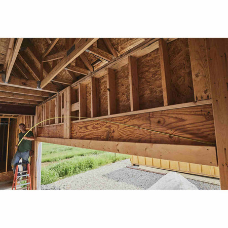 An action shot of a professional contractor using the Stanley 30 ft FATMAX tape measure on a residential framing jobsite, demonstrating the 14-foot reach capability while measuring a large wooden header solo.