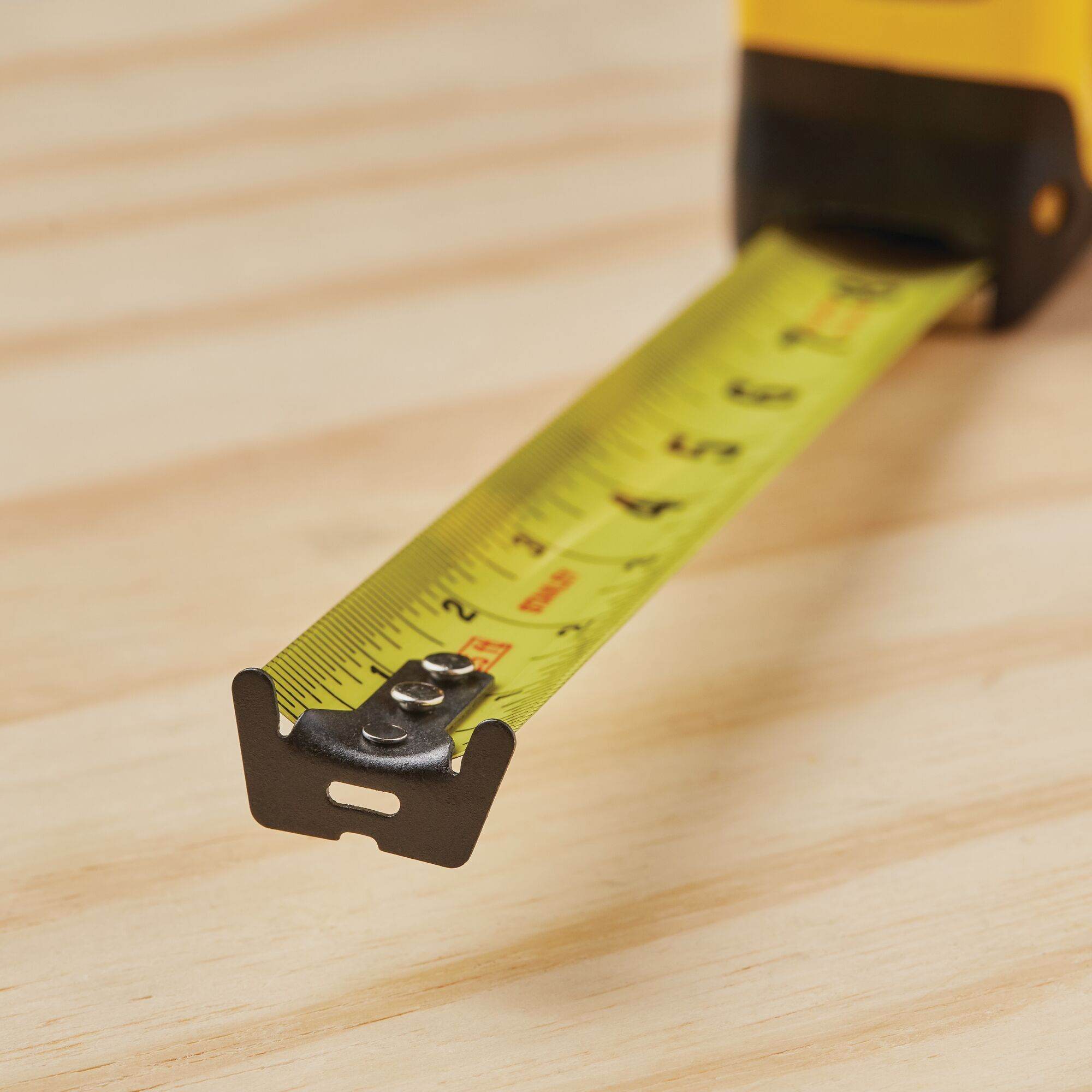 A detailed close-up view of the 1-1/4 inch wide yellow steel blade of the Stanley FATMAX tape measure extended over a wood surface, showcasing the durable multi-catch hook and fractional measurement markings.