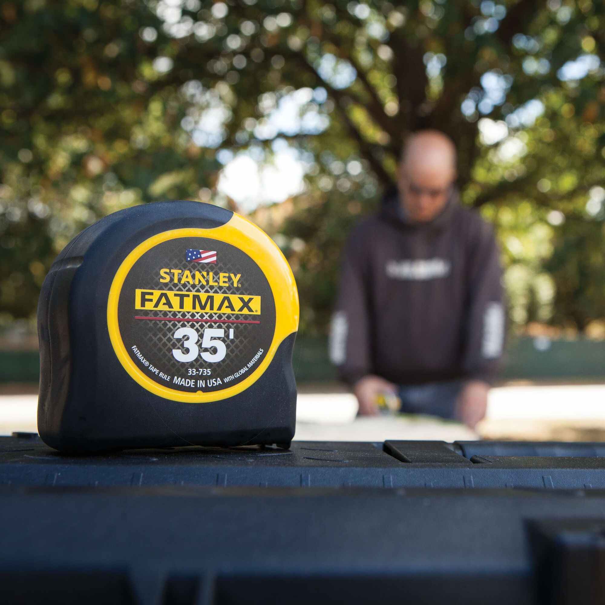 A Stanley 35 foot FATMAX tape measure resting on a black surface in the foreground with a professional contractor working on a project in the blurred background.