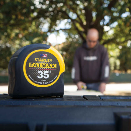 A Stanley 35 foot FATMAX tape measure resting on a black surface in the foreground with a professional contractor working on a project in the blurred background.