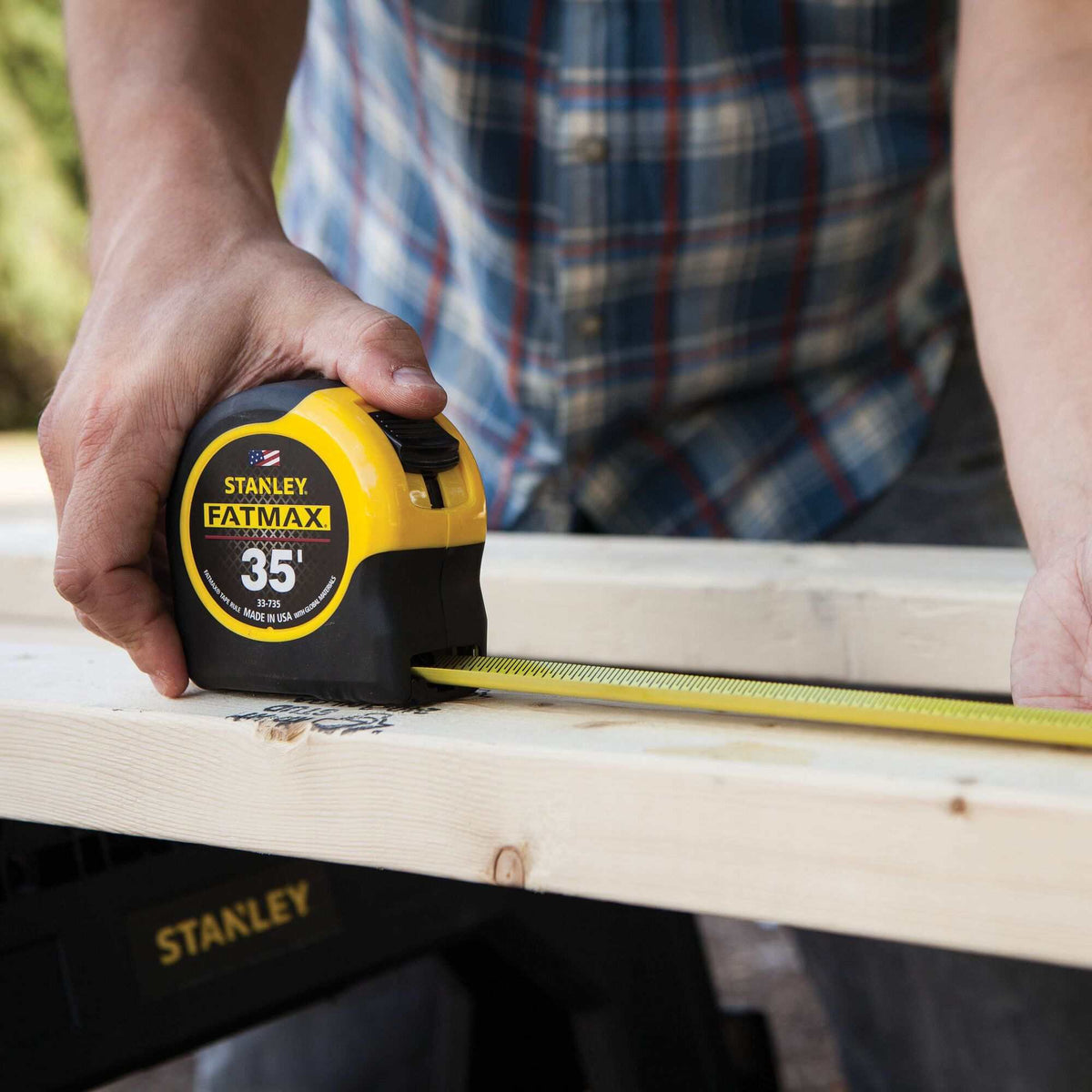 Close-up of a worker's hands using the Stanley FATMAX 35 foot tape measure to take an accurate measurement on a wooden 2x4 board at a construction site.