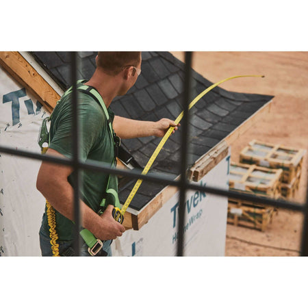 Over-the-shoulder view through a window frame of a contractor using the high-visibility yellow blade of a Stanley FATMAX tape measure to layout structural measurements on a house.