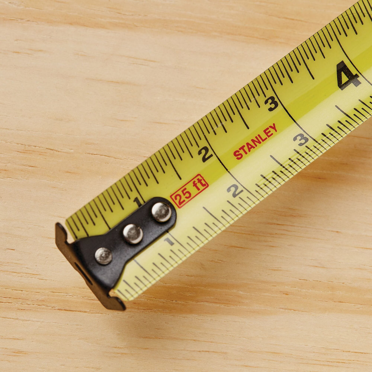 A detailed top-down product shot of the initial segment of the yellow Stanley steel measuring blade, highlighting the prominent red 25 ft length stamp, the multi-rivet black reinforcement bracket, and clear black fractional numbers.