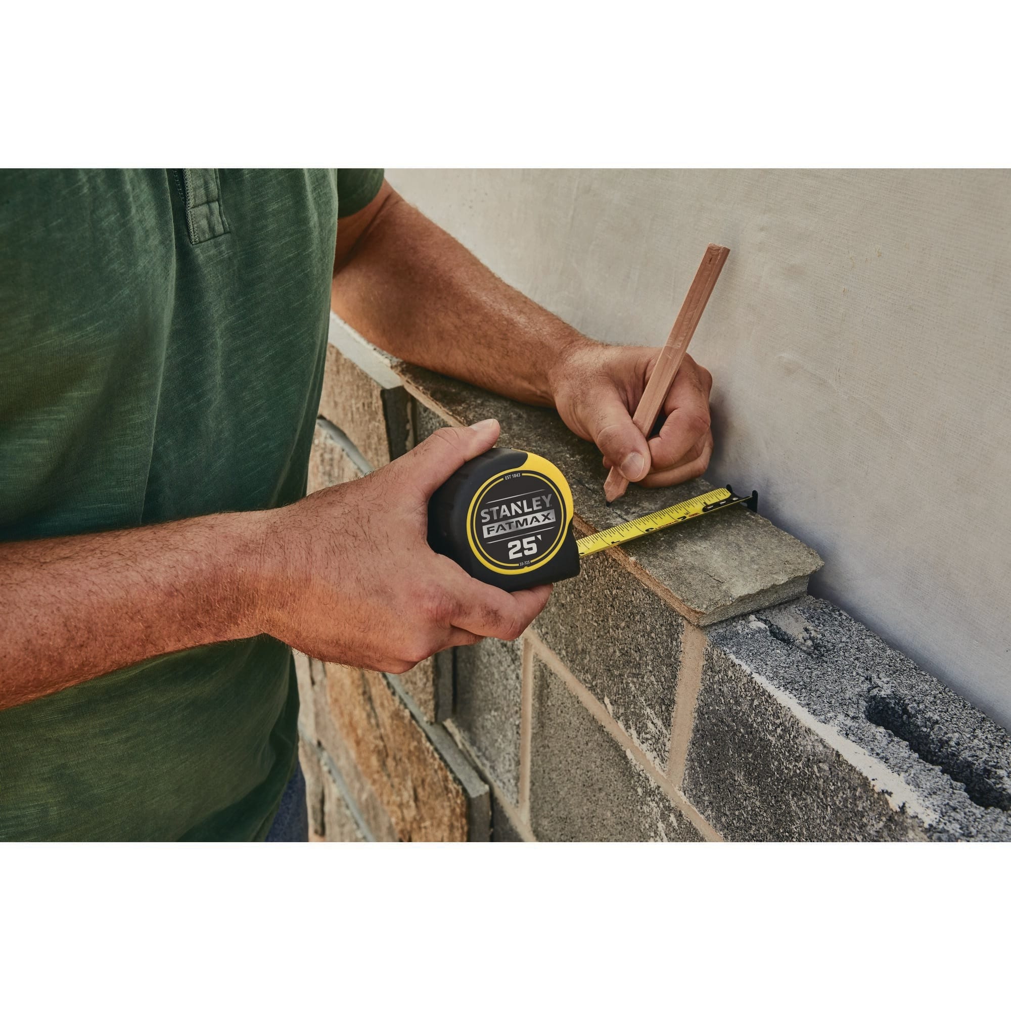 An outdoor jobsite stone masonry application photograph showing a tradesman using the compact yellow and black Stanley FatMax 25-foot rule and a timber pencil to mark layout lines along a rough concrete block and stone veneer wall.