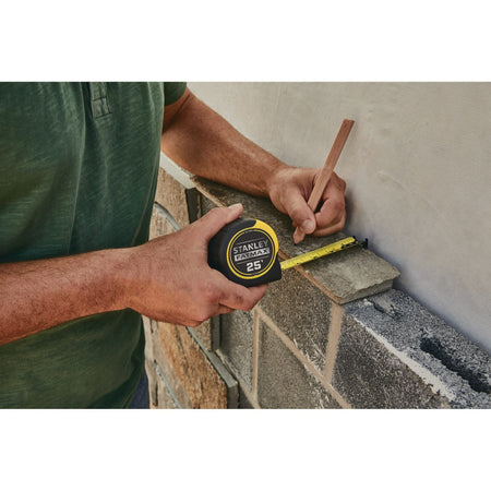 An outdoor jobsite stone masonry application photograph showing a tradesman using the compact yellow and black Stanley FatMax 25-foot rule and a timber pencil to mark layout lines along a rough concrete block and stone veneer wall.