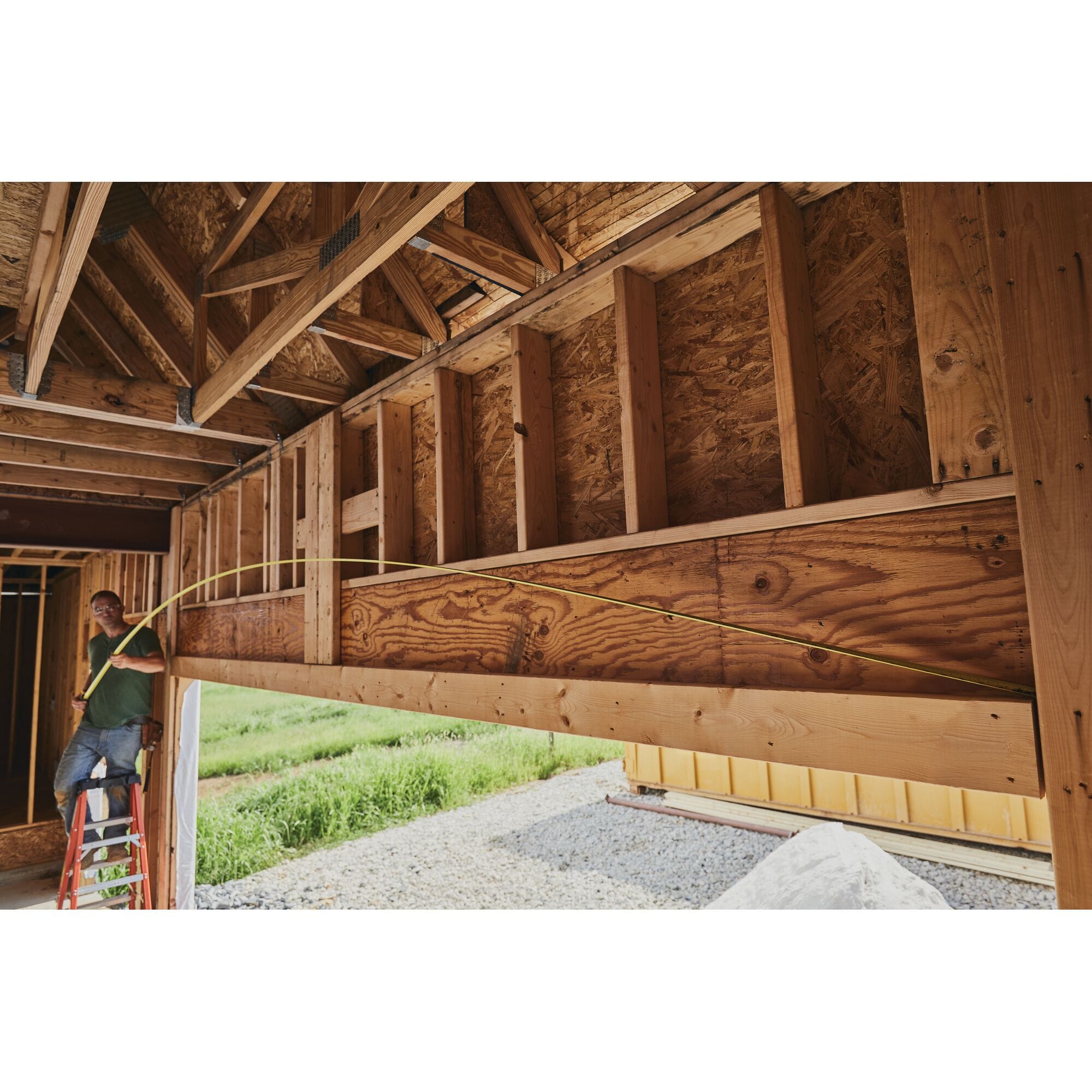 A real-world commercial construction application photograph showing a contractor standing on a ladder inside a home framework, testing the 14-foot total reach capacity by extending the yellow steel blade of the FatMax tape horizontally across a massive wood ceiling header.