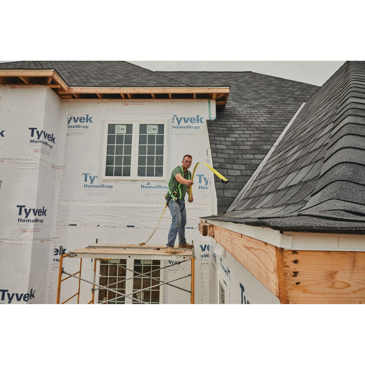 A wide-angle outdoor lifestyle application shot showing a roofer wearing a safety body harness standing on scaffolding, extending the yellow steel blade of the dynamic Stanley FatMax tape measure high into the air to verify structural alignment against an asphalt shingle roof.