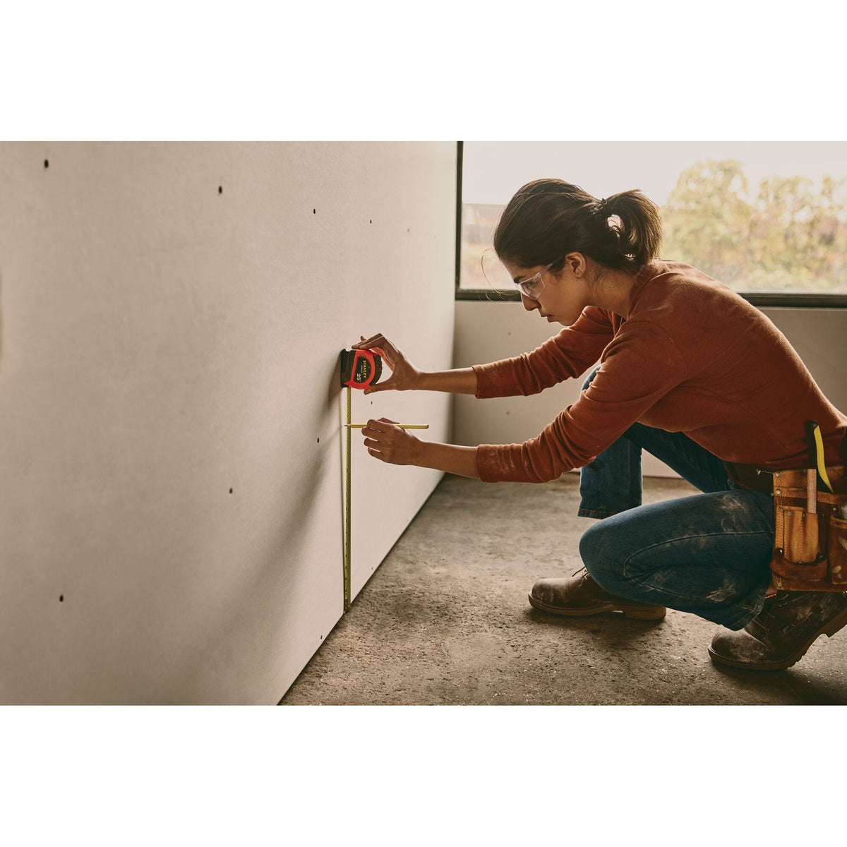 An indoor construction lifestyle application shot showing a female professional wearing safety glasses crouching while using the high-visibility orange Stanley LeverLock 25-foot tape measure and a pencil to mark vertical dimensions on a drywall panel board.