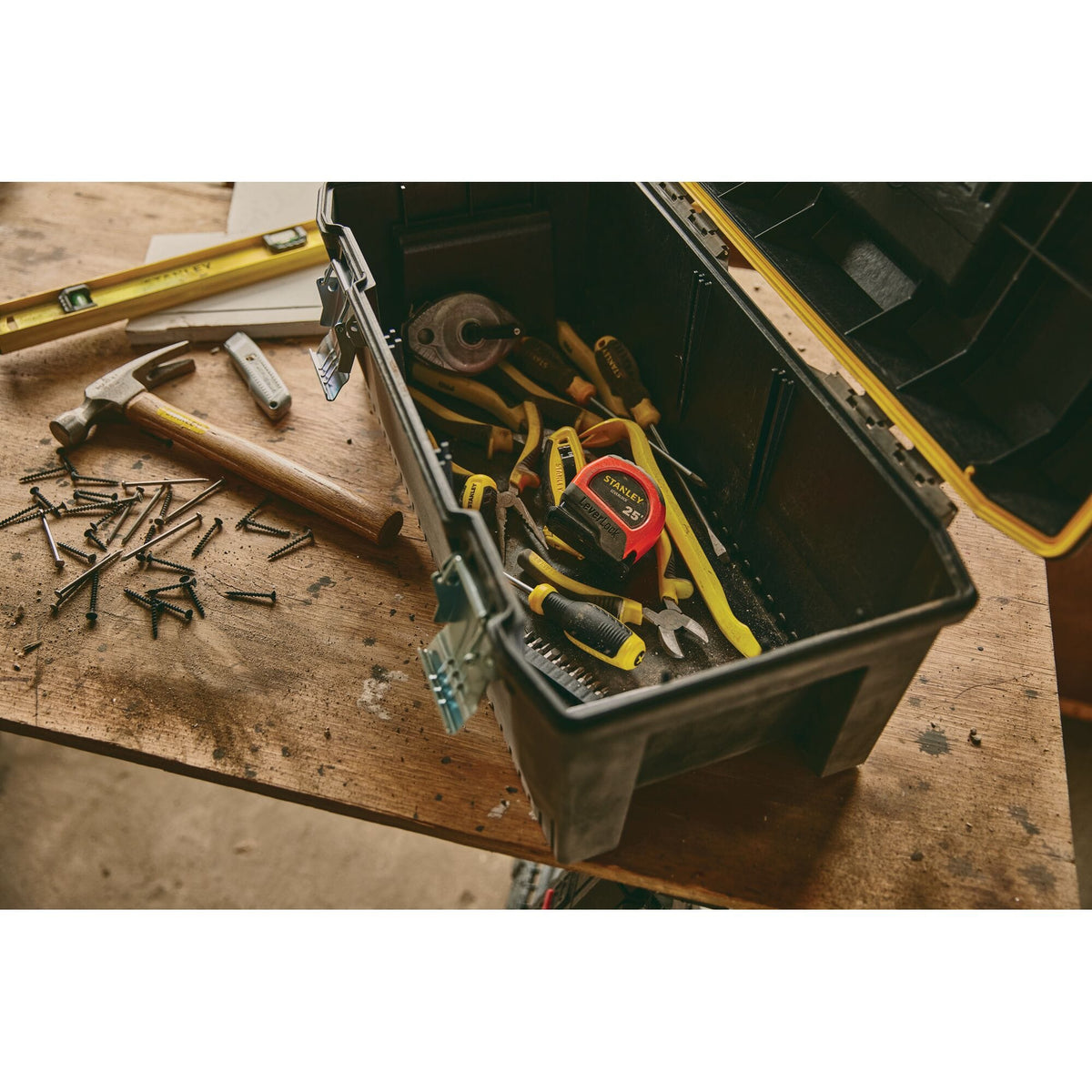 A real-world e-commerce lifestyle photograph showing an open black plastic tool box resting on a wooden workbench, showcasing the high-visibility orange Stanley LeverLock 25' tape measure easily identifiable among various hand tools, screws, a hammer, and a level.