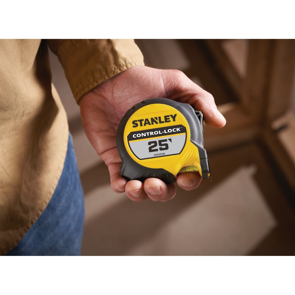 A close-up lifestyle product photograph of the compact Stanley Control-Lock 25' tape measure case resting flat in the palm of a tradesperson's hand inside a residential wood framing environment.