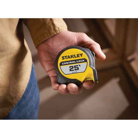 A close-up lifestyle product photograph of the compact Stanley Control-Lock 25' tape measure case resting flat in the palm of a tradesperson's hand inside a residential wood framing environment.