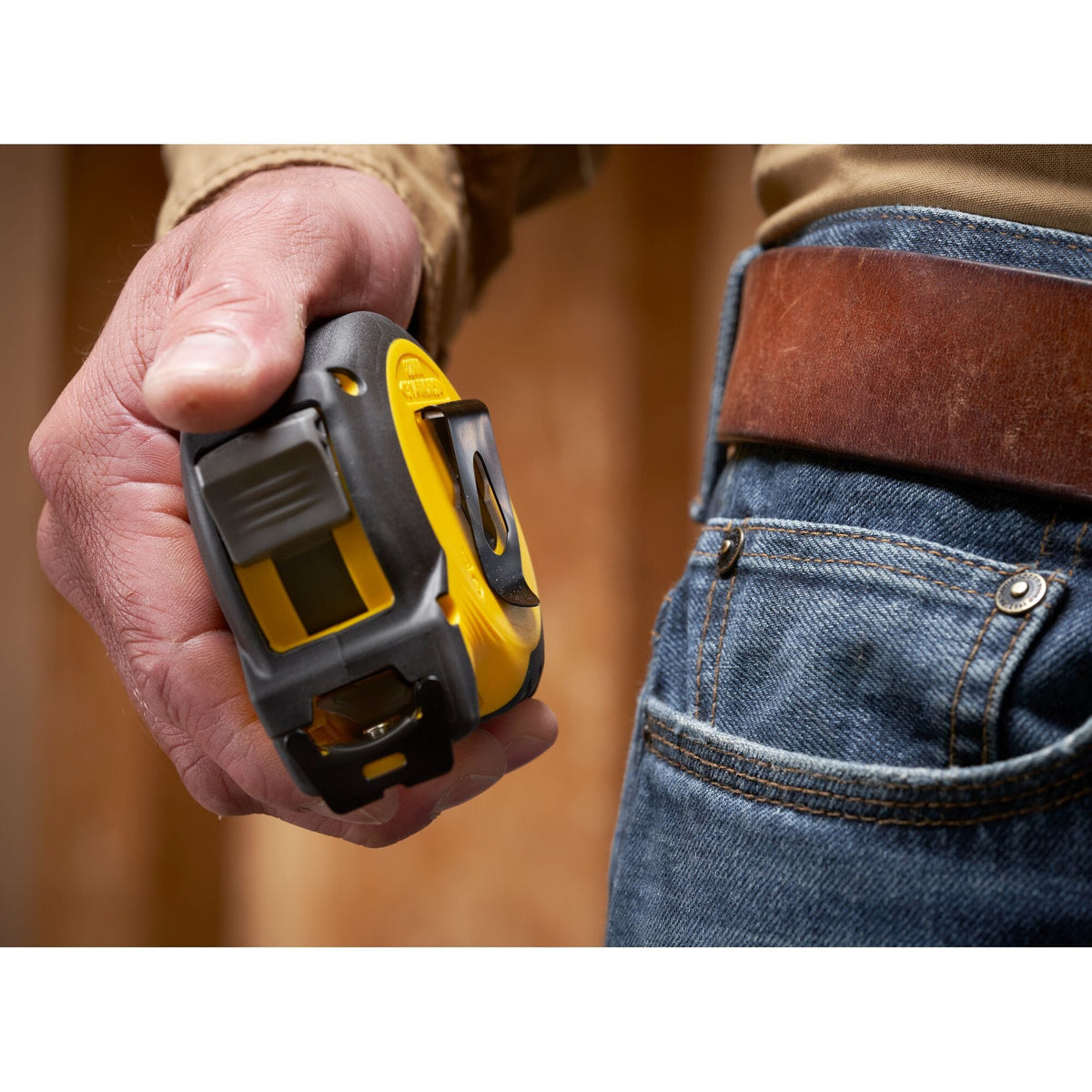 A close-up real-world photo of a worker holding the black and yellow Stanley measuring tape case, demonstrating the reverse-mounted black steel pocket clip positioning near a leather tool belt and blue jeans pocket.