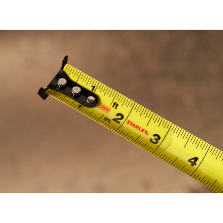 A detailed macro view of the yellow steel ribbon of the Stanley Control-Lock 25ft rule, showcasing the three structural rivets on the black multi-catch dual hook tip and clear fractional marking lines against a blurred background.