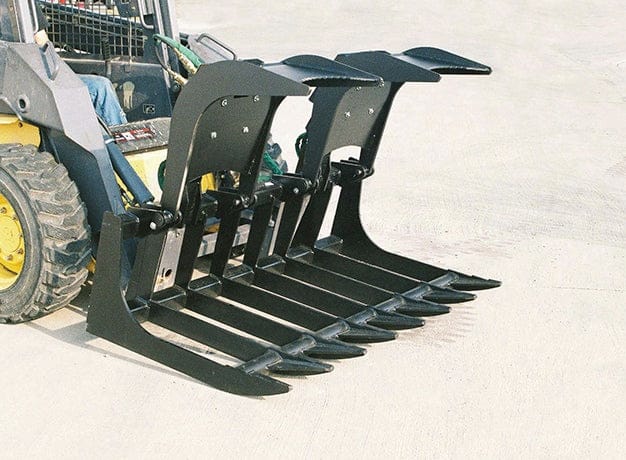 High angle view of the root grapple attached to a skid steer loader resting on a concrete surface, showing the dual independent hydraulic clamps in the open position and the protected hydraulic cylinder housings.