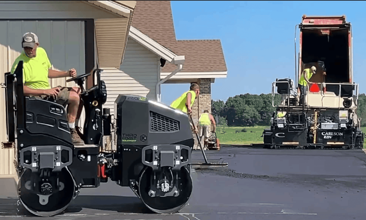 An industrial paving crew uses the Third Coast Equipment RA12H vibratory tandem roller to finish a residential driveway, demonstrating the machine's smooth finishing capabilities alongside a large asphalt paver.