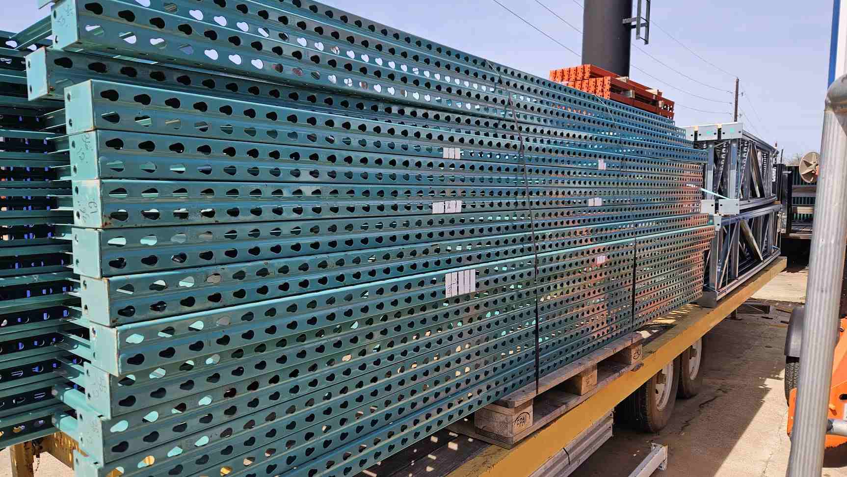A wide side-profile view of a large stack of used like new green teardrop pallet rack uprights stored on a transport trailer, highlighting the 12-foot height and 36-inch depth with reinforced steel bracing for industrial warehouse storage.