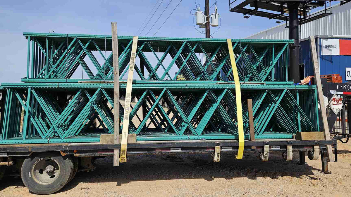 A wide side-profile view of multiple used like new green teardrop pallet rack uprights stacked and secured with yellow straps on a flatbed transport trailer, showcasing the 20-foot height and 42-inch depth for industrial warehouse storage.