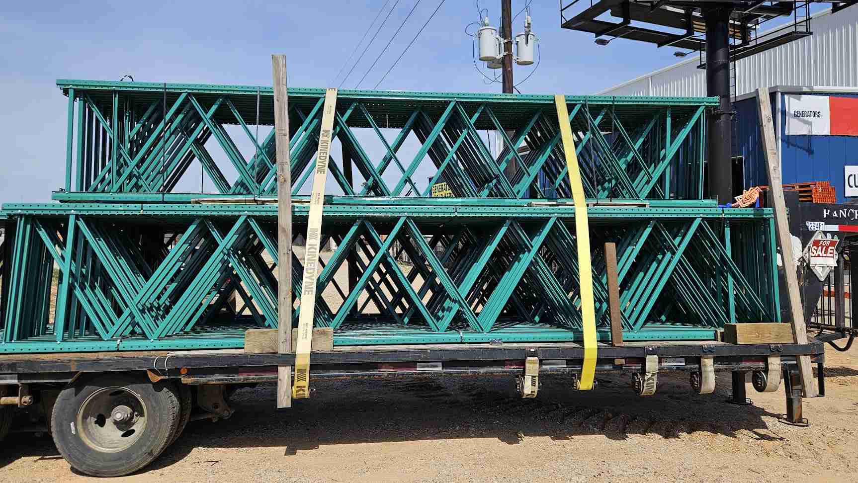 A wide side-profile view of multiple used like new green teardrop pallet rack uprights stacked and secured with yellow straps on a flatbed transport trailer, showcasing the 20-foot height and 42-inch depth for industrial warehouse storage.