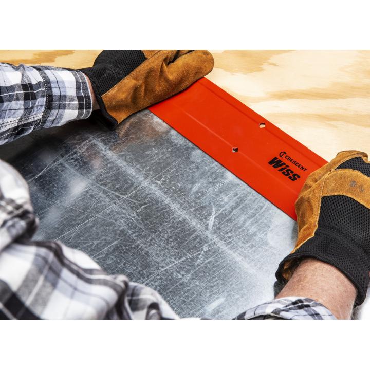 Imagine feeling a flat, rectangular metal tool with raised letters, held firmly by two hands wearing textured gloves. The tool is being pressed down onto a cool, smooth sheet of metal, bending it along the tool's edge.