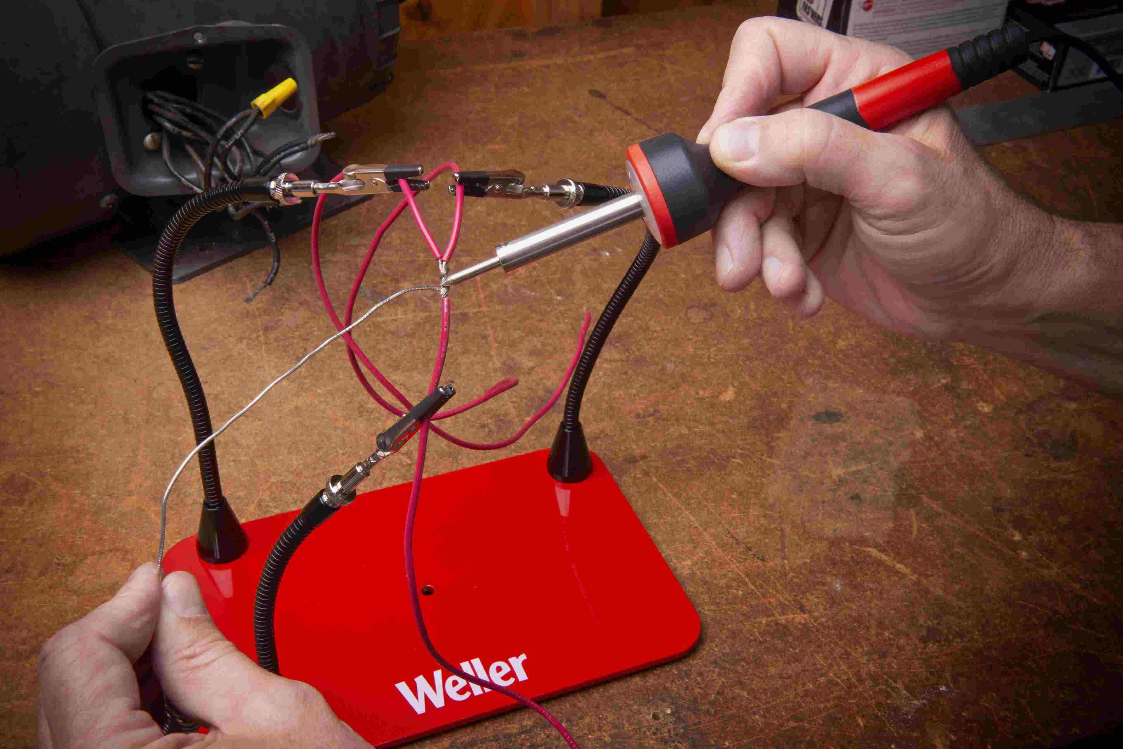 Person soldering wires held by Weller helping hands tool with red base and flexible arms.